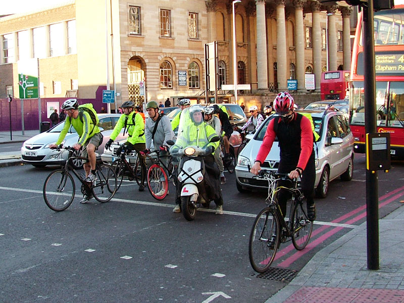 Radfahrer in London warten an der Ampel, von Autos umgeben. Die Radfahrer tragen alle Warnwesten und Helme.
