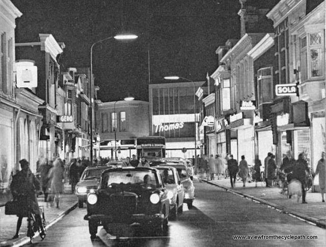 Ein Foto von Assen in der 1960er Jahren. Fußgänger sind an den Rand gedrängt, während ein einsamer Radfahrer neben den Autos an der Ampel wartet.